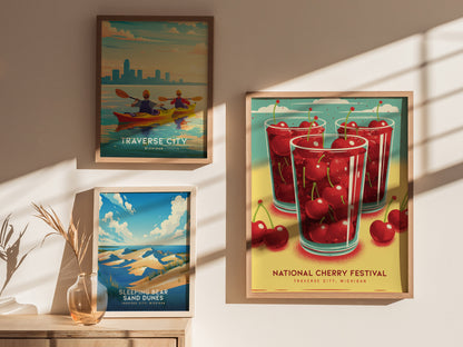 Framed posters on a sunlit wall depict Traverse City, Michigan with vibrant scenes: kayakers, Sleeping Bear Sand Dunes, and the National Cherry Festival featuring glasses filled with cherries.