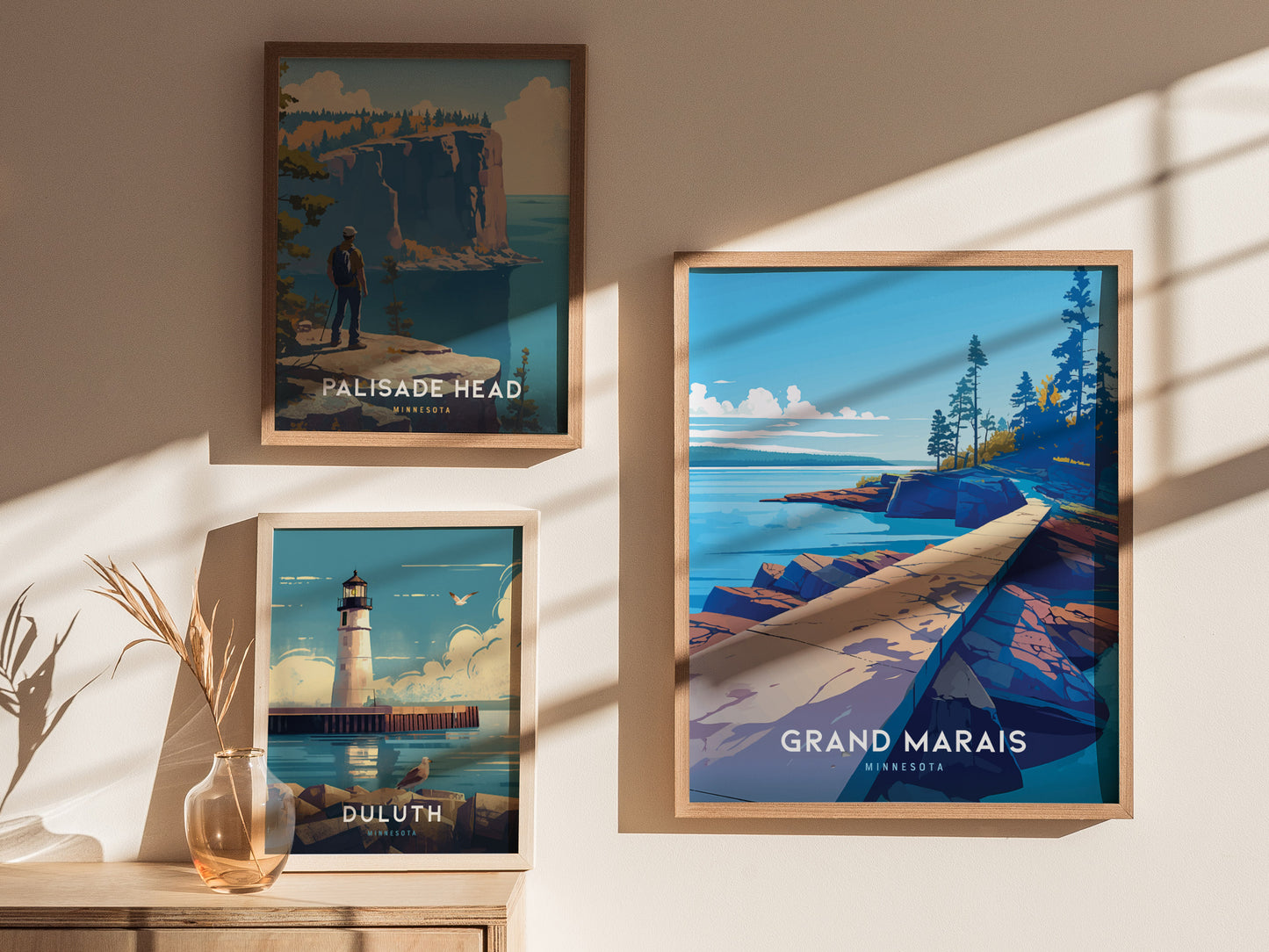 Three framed posters hang on a wall, depicting scenic landscapes of Minnesota: Palisade Head, a lighthouse in Duluth, and the shoreline of Grand Marais. Shadows and a vase add warmth.