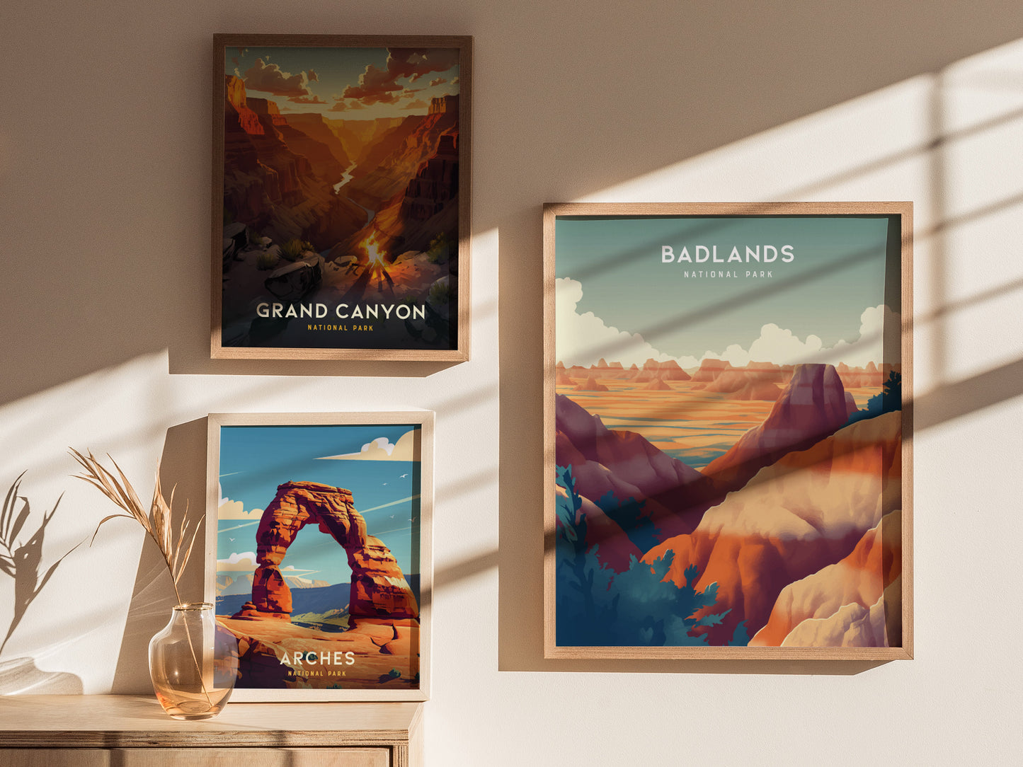 Three framed posters depict landscapes: Grand Canyon, Arches, and Badlands National Park. They are displayed on a wall with sunlight casting shadows, beside a vase with dried plants.