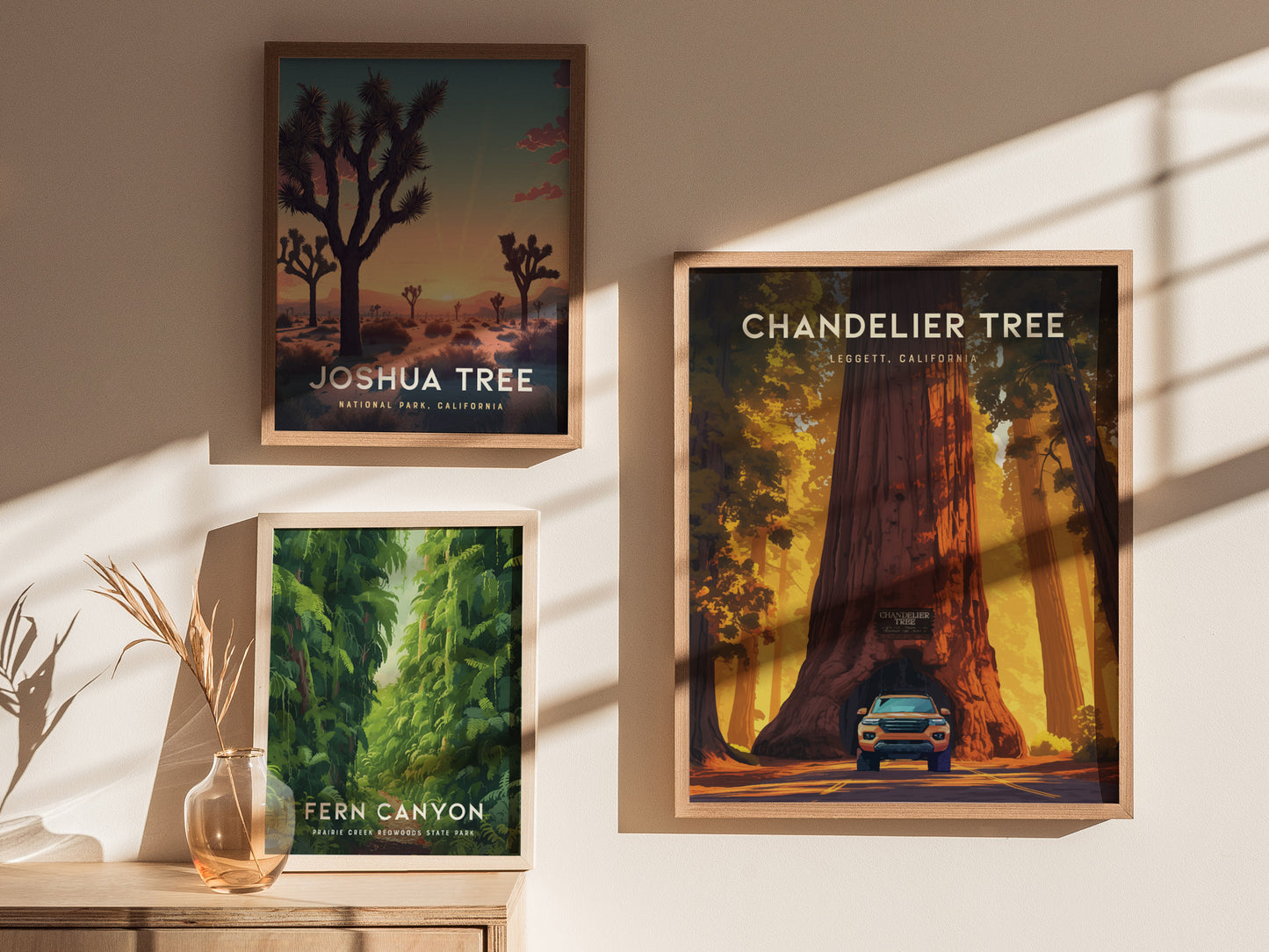 Three framed posters on a wall depict Joshua Tree, Chandelier Tree with a car, and Fern Canyon. Sunlight casts shadows, and a vase with dried plants is on a wooden.