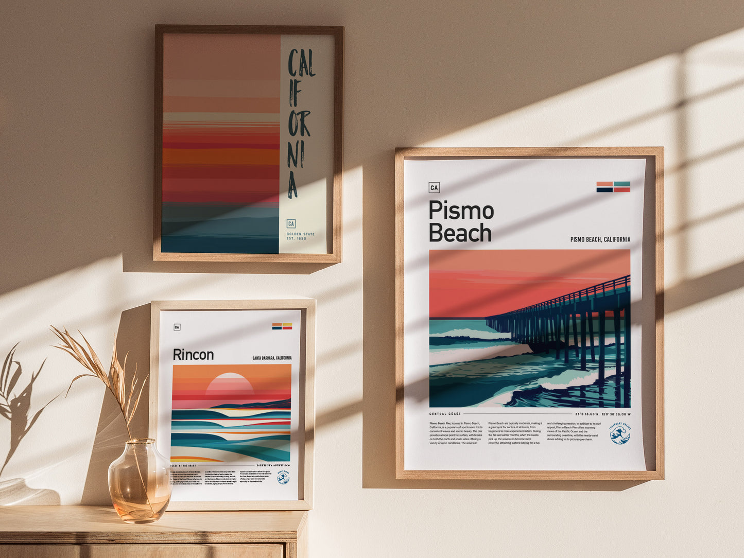 Three framed posters hang on a wall. The largest features a stylized Pismo Beach pier at sunset. Nearby are two smaller posters, one with abstract ocean waves and another with.