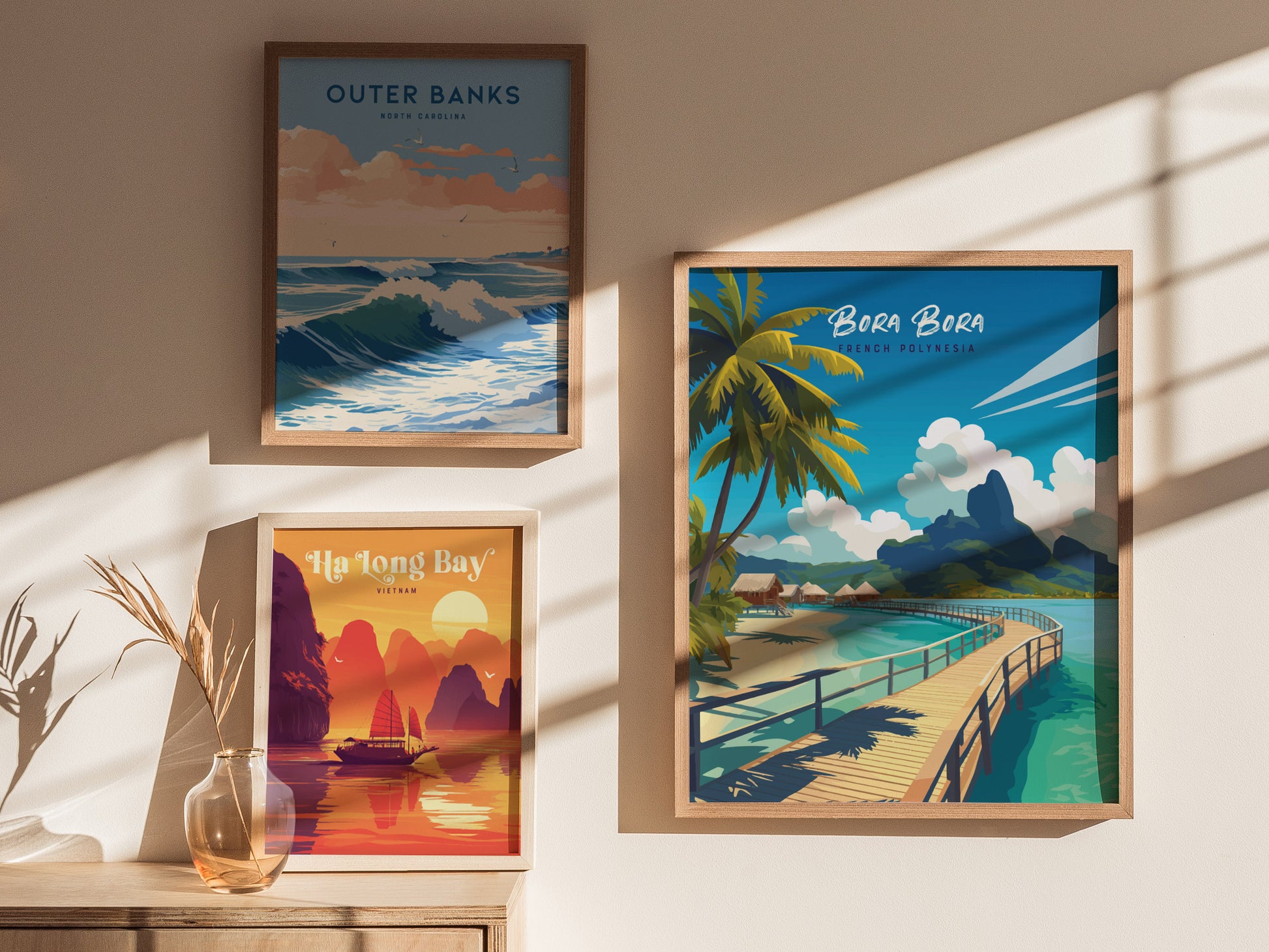 Three framed travel prints hang on a sunlit wall, depicting Outer Banks, Bora Bora, and Ha Long Bay. A small vase with dried plants sits below the artwork.