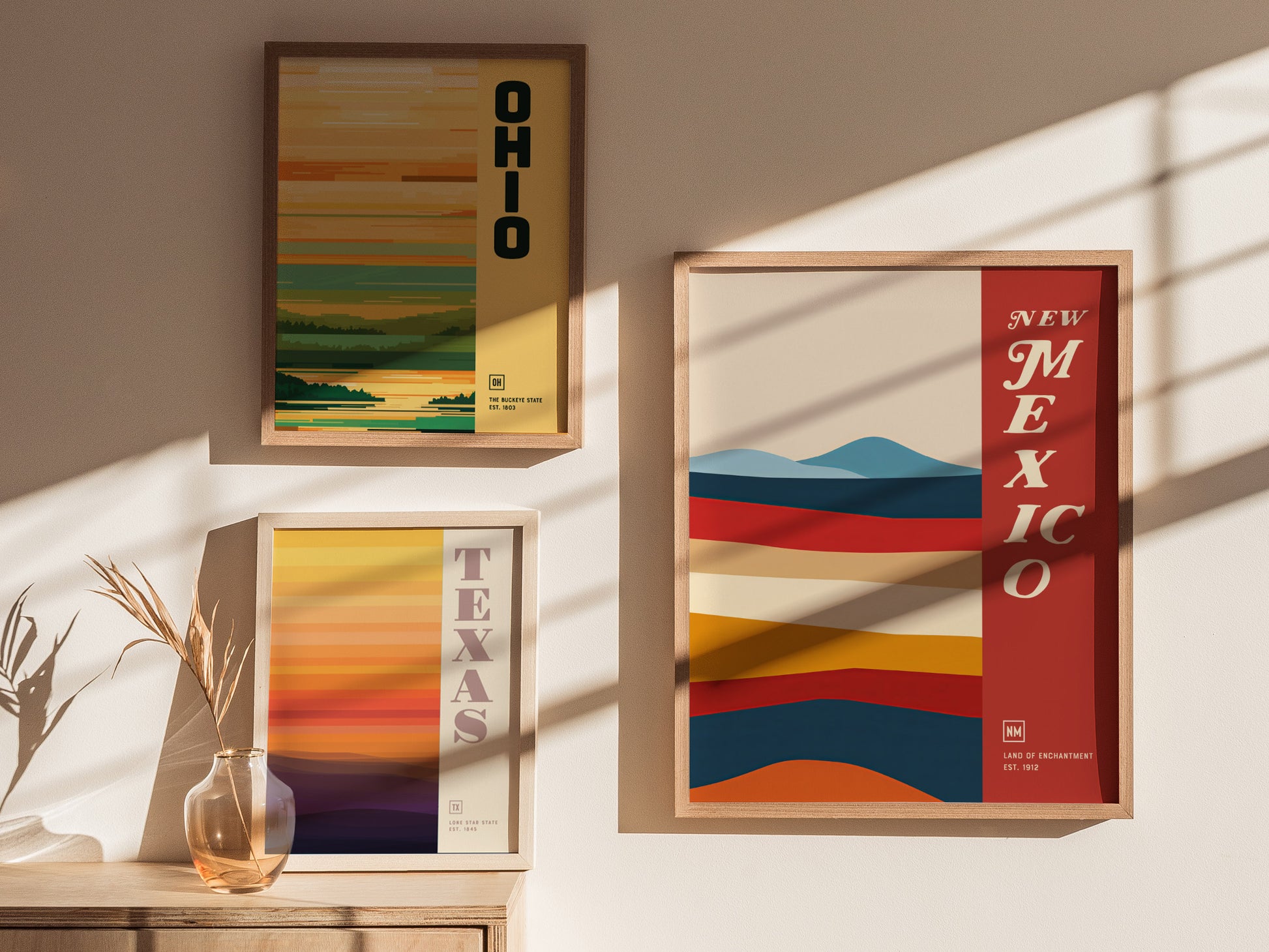 Three framed posters are displayed on a sunlit wall, featuring abstract designs and the names Ohio, Texas, and New Mexico. A small vase with dried plants sits below.