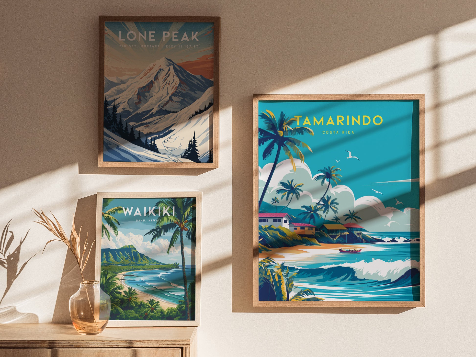 Three framed posters hang on a sunlit wall, featuring vibrant beach scenes of Tamarindo, Lone Peak, and Waikiki, with a small vase and dried plant on a wooden surface.