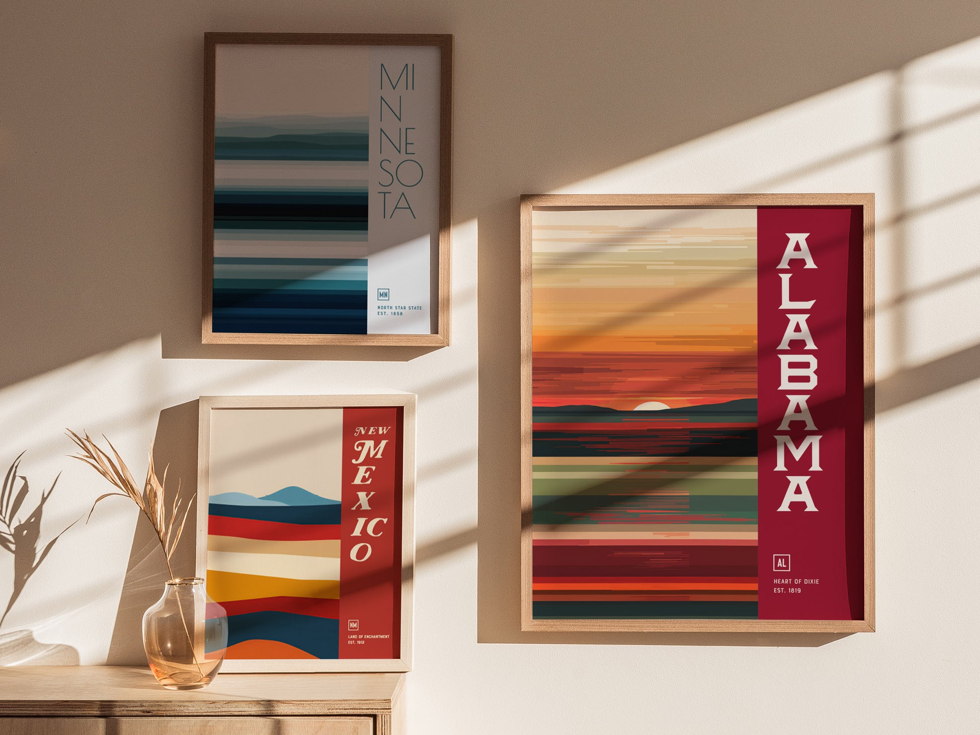 Three framed minimalist posters featuring abstract designs for Minnesota, Alabama, and New Mexico are displayed on a wall with warm lighting and shadows.