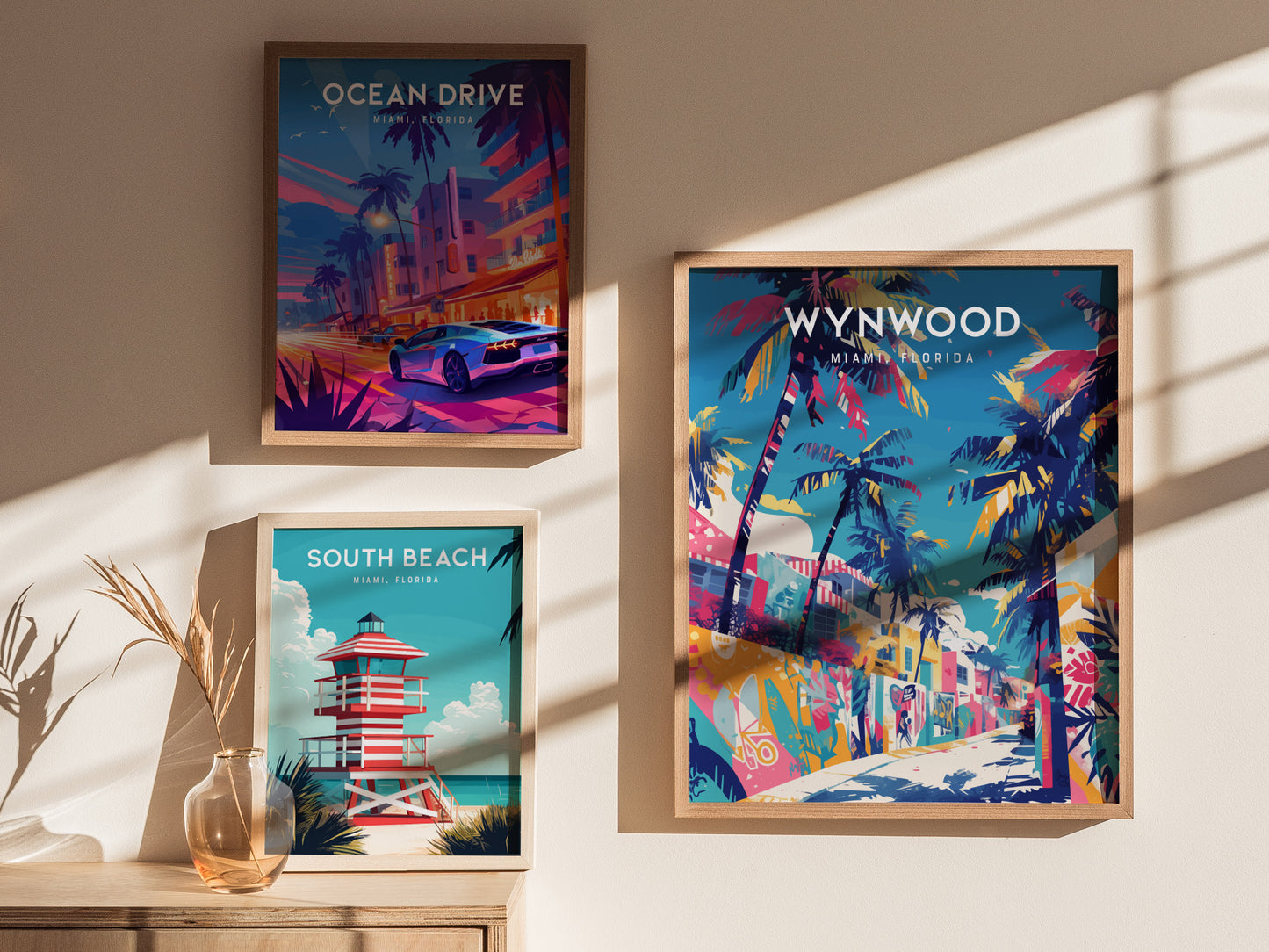 Three framed posters are displayed on a sunlit wall. The top left poster features Ocean Drive with a vibrant night scene, the bottom left shows a South Beach lifeguard tower, and the right poster depicts colorful Wynwood street art.