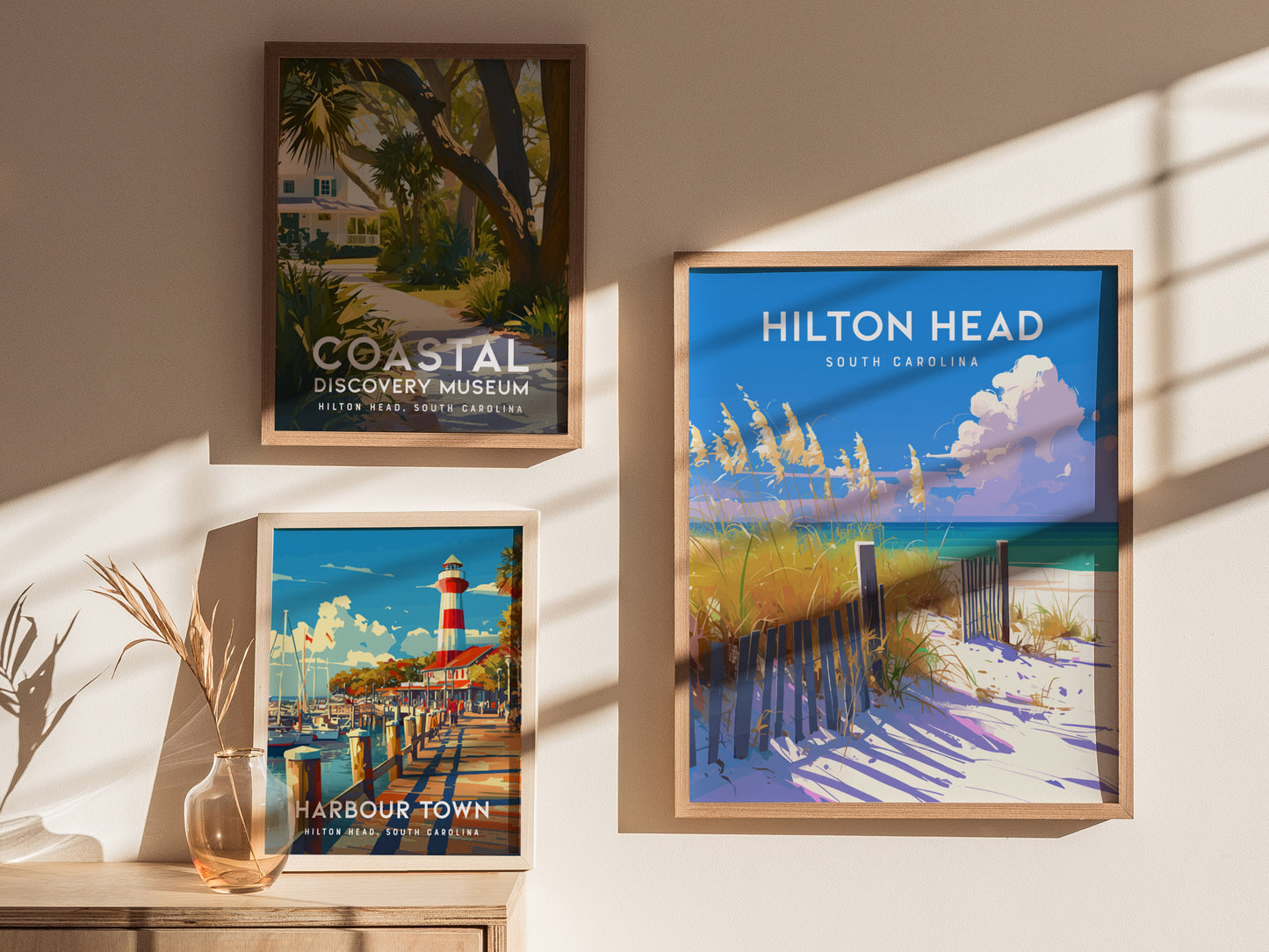 Three framed posters are displayed on a wall, showcasing coastal scenes. The top left poster features a museum surrounded by lush greenery, the bottom left shows a harbor with a lighthouse, and the right poster depicts a sandy beach.