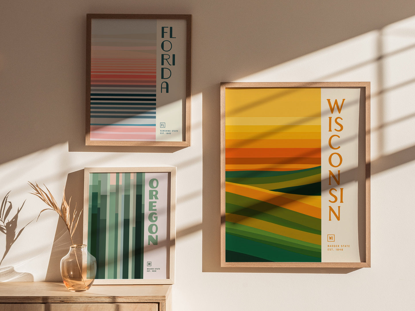 Three framed minimalist posters are displayed on a wall, each representing Florida, Oregon, and Wisconsin with abstract patterns in shades of green, gold, and pink.
