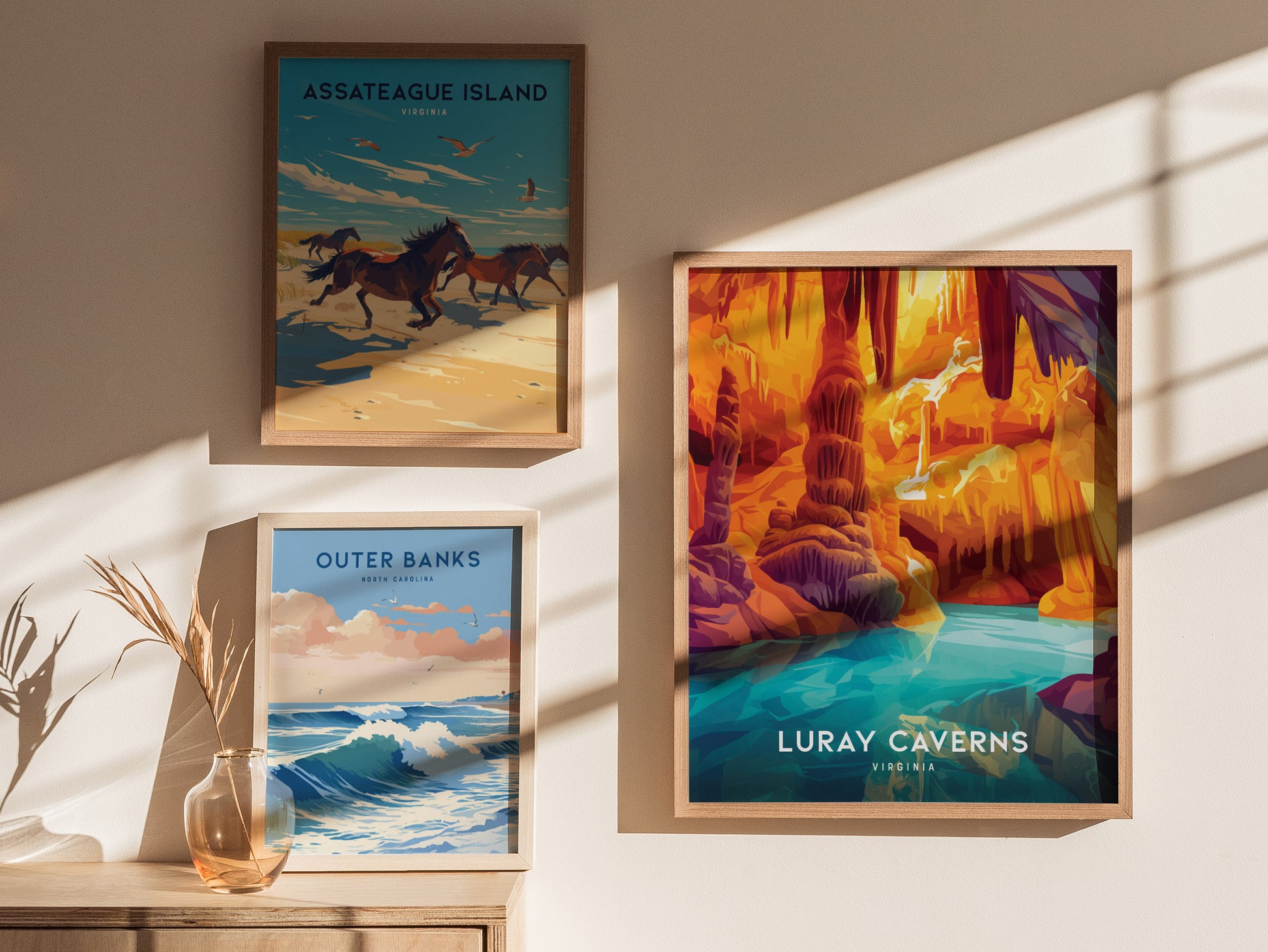 Three framed posters are displayed on a sunlit wall, featuring Luray Caverns, Assateague Island with horses, and Outer Banks with ocean waves, complemented by a glass vase with dried plants.