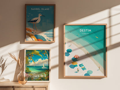 Three framed posters are displayed on a sunlit wall, featuring Sanibel Island, Destin, and Islamorada with vibrant beach and ocean scenes. Shadows and a decorative vase add depth.