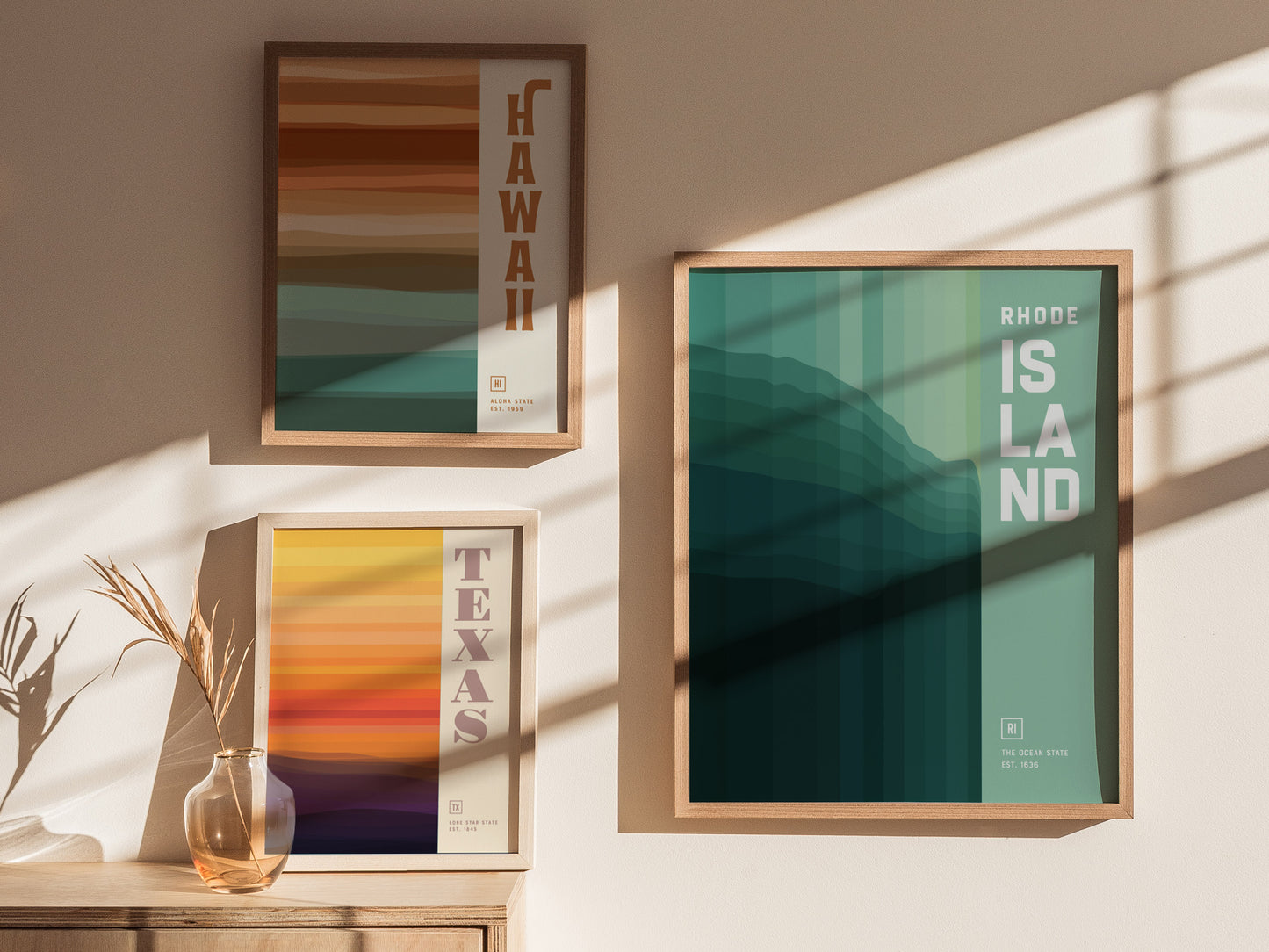 Three framed posters on a sunlit wall depict abstract designs representing Hawaii, Texas, and Rhode Island, featuring layered colors and bold text. A vase with dried plants sits nearby.