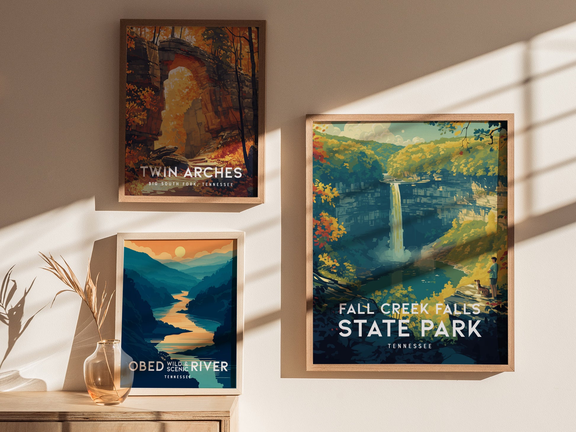 Three framed posters are displayed on a wall, featuring scenic illustrations of Twin Arches, Obed Wild & Scenic River, and Fall Creek Falls State Park in Tennessee. Sunlight casts shadows.