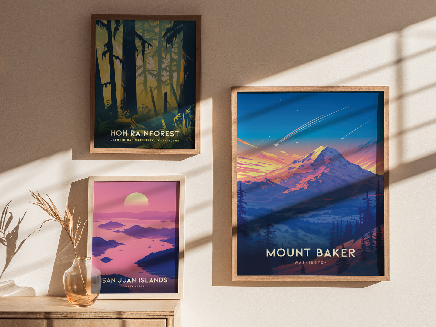 Three framed posters are displayed on a sunlit wall: Hoh Rainforest, San Juan Islands, and Mount Baker. A glass vase with dried plants sits on a wooden surface.