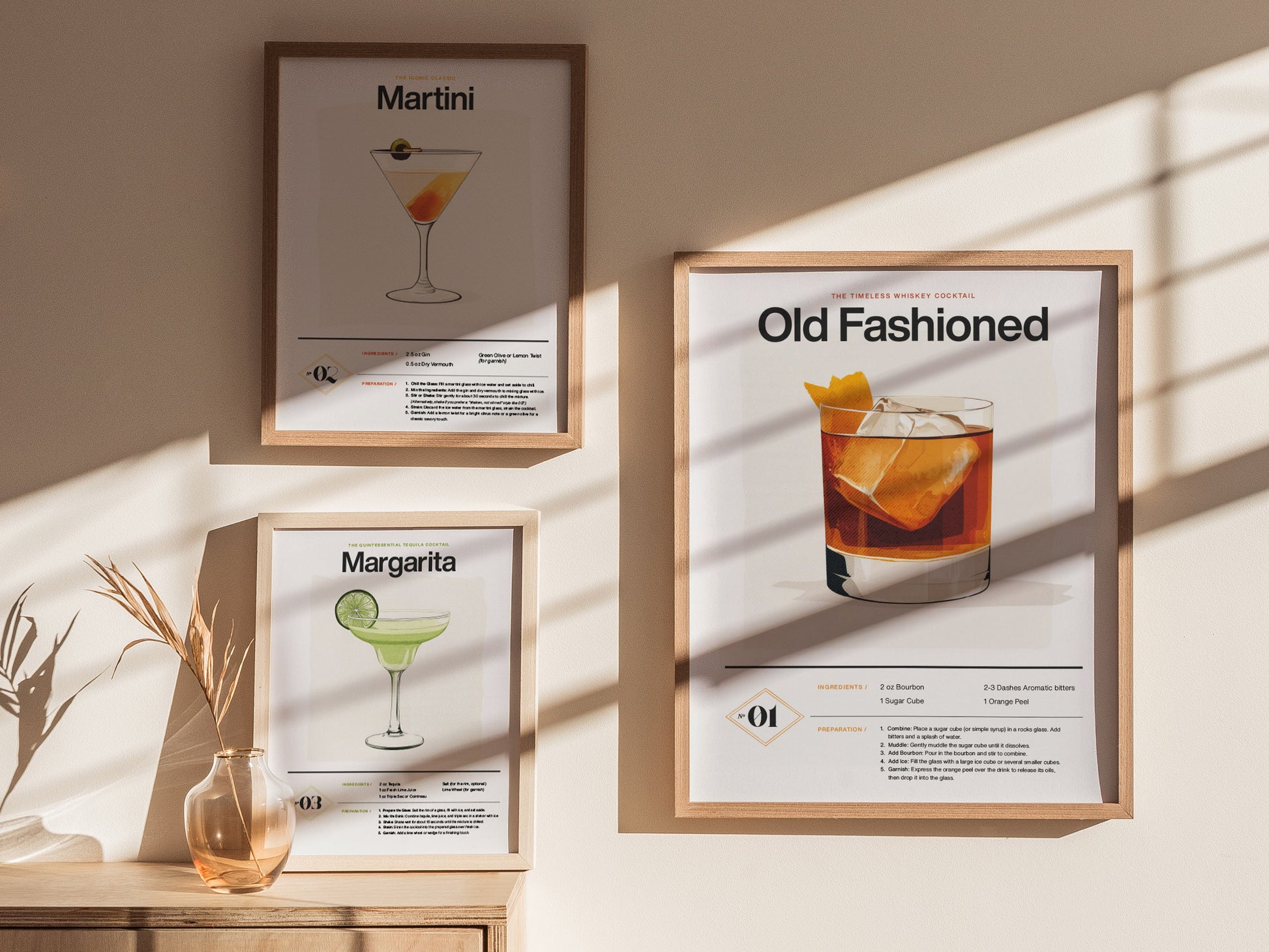 Three framed cocktail posters on a wall depict Martini, Margarita, and Old Fashioned recipes. Sunlight casts shadows, and a vase with dried grass is on a wooden surface.