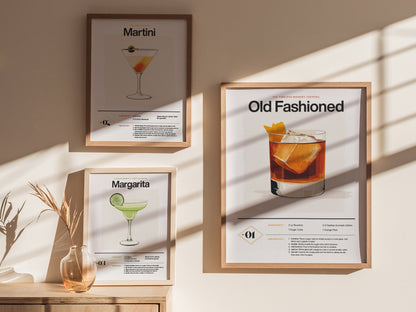 Three framed cocktail posters on a wall depict Martini, Margarita, and Old Fashioned recipes. Sunlight casts shadows, and a vase with dried grass is on a wooden surface.