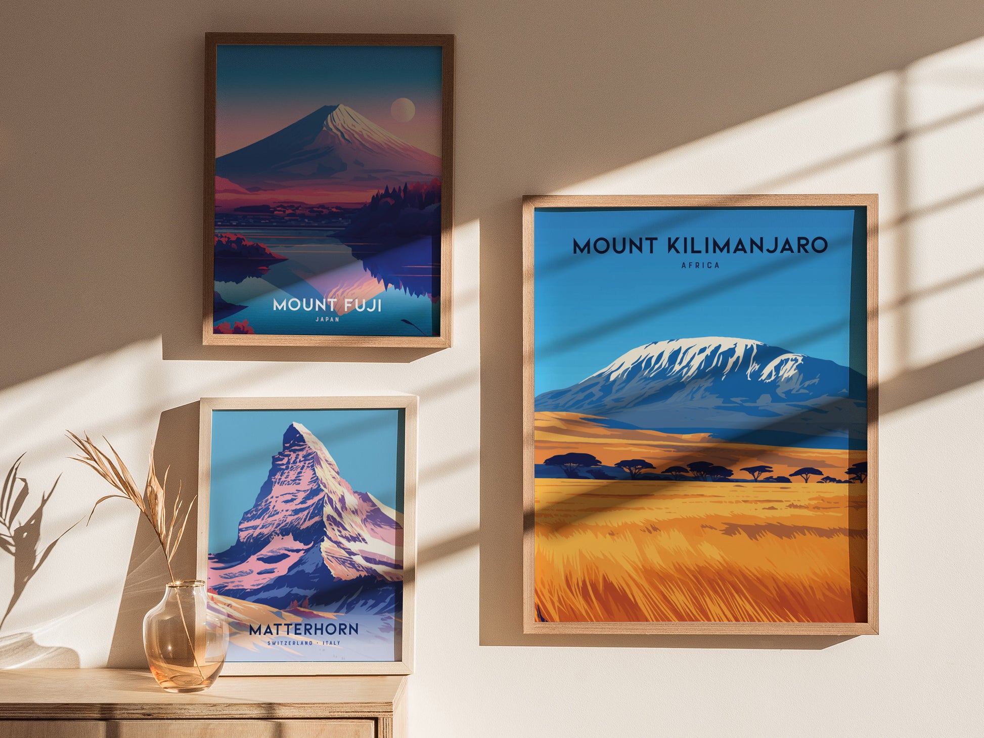 Three framed posters on a sunlit wall depict Mount Fuji, Mount Kilimanjaro, and the Matterhorn, each showcasing vibrant landscapes and iconic peaks. A decorative vase with dry plants sits below.