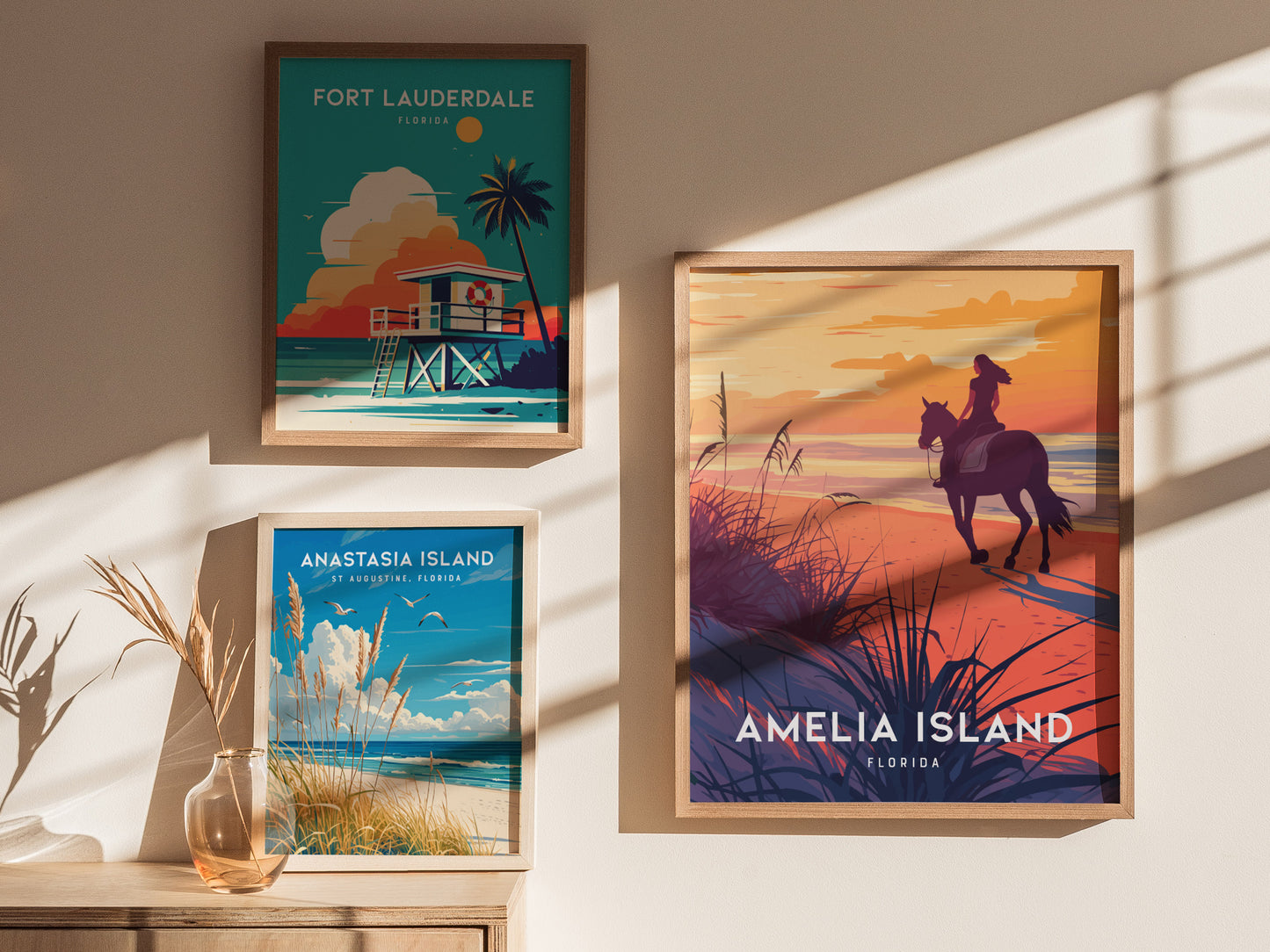 Three framed posters are displayed on a sunlit wall, featuring scenes from Fort Lauderdale, Anastasia Island, and Amelia Island, each depicting vibrant beach landscapes and coastal elements.