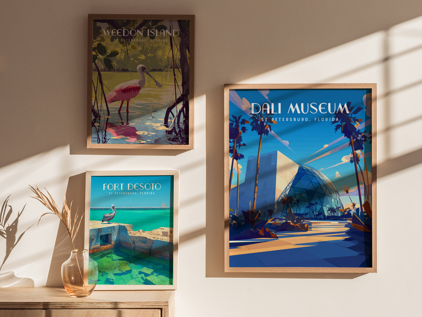 Framed posters on a sunlit wall featuring Weedon Island, Fort Desoto, and the Dali Museum in vibrant, artistic styles. A glass vase with dried grass decorates a wooden table.