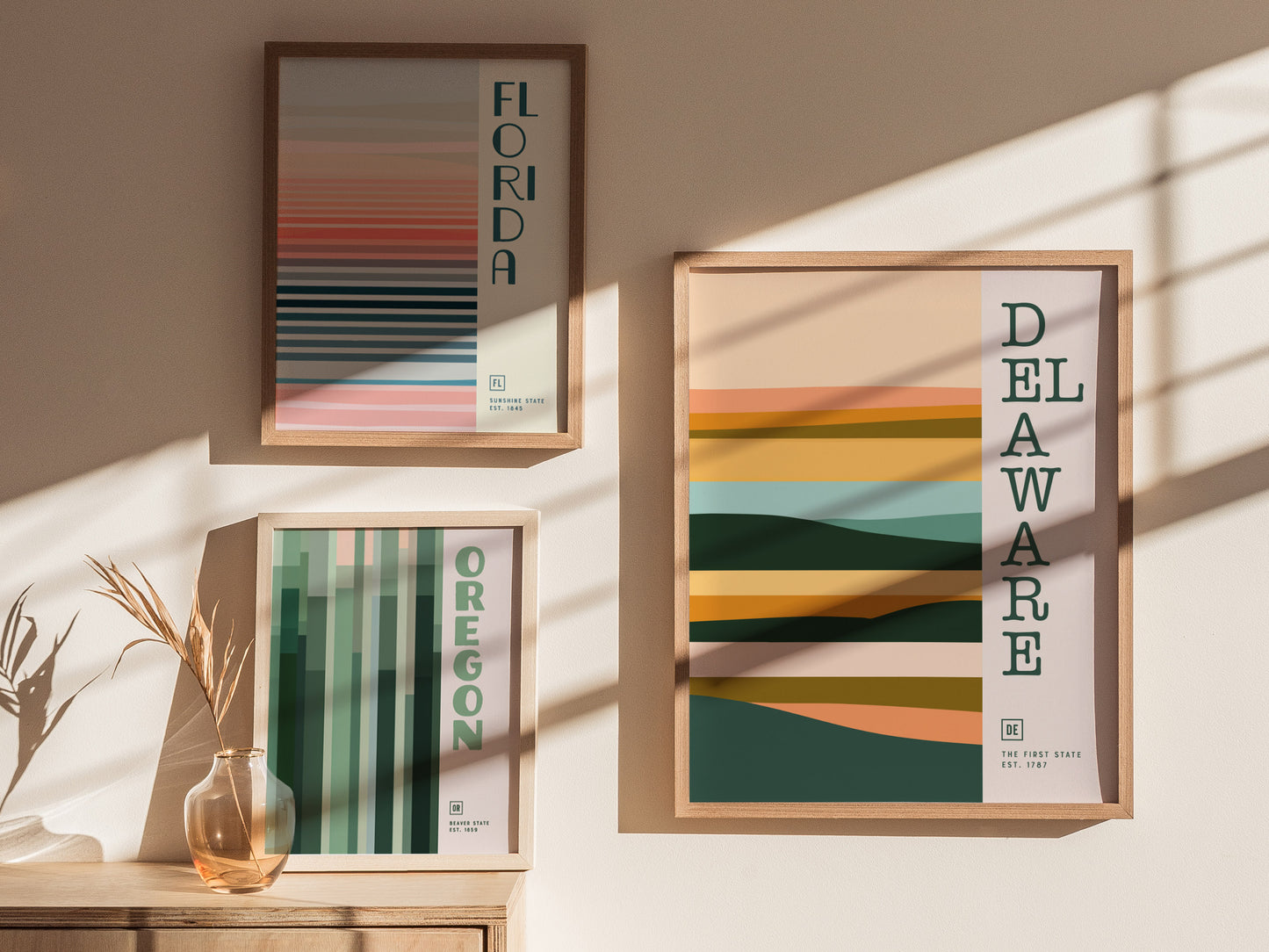 Three framed minimalist posters featuring abstract designs of Florida, Delaware, and Oregon hang on a sunlit wall. A glass vase with dry foliage sits on a wooden surface.