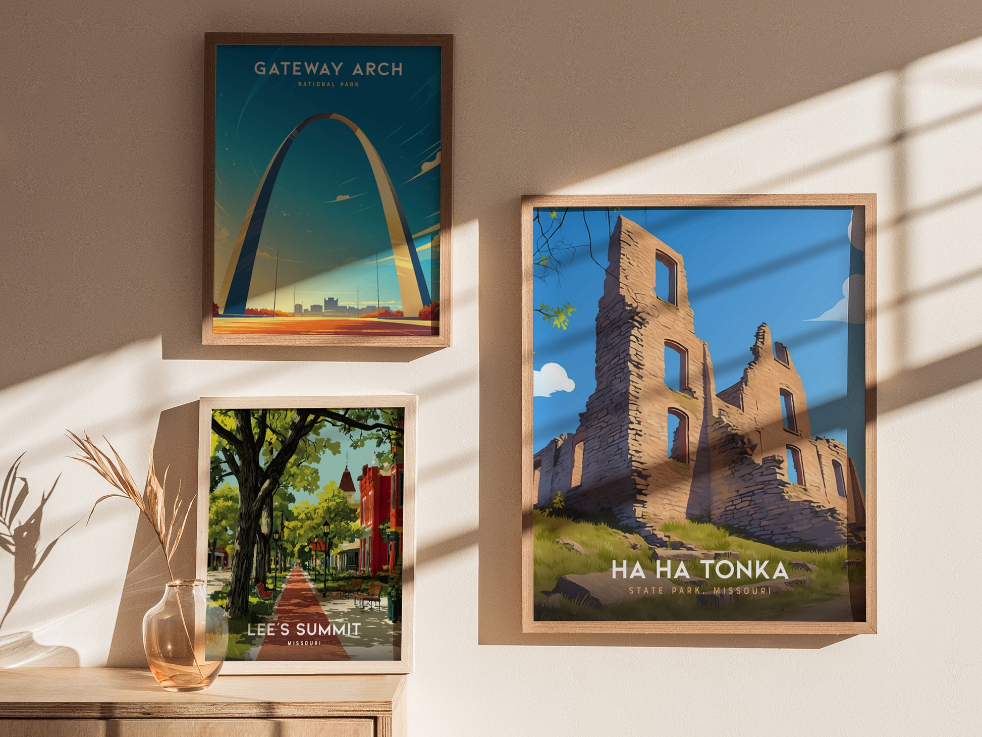 Three framed posters hang on a sunlit wall, depicting Gateway Arch, a tree-lined street in Lees Summit, and the ruins of Ha Ha Tonka State Park.