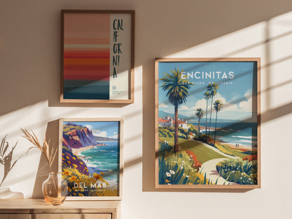 Framed posters of Encinitas, Del Mar, and California hang on a sunlit wall, featuring coastal scenes with palm trees and cliffs. A vase with dried plants sits below.