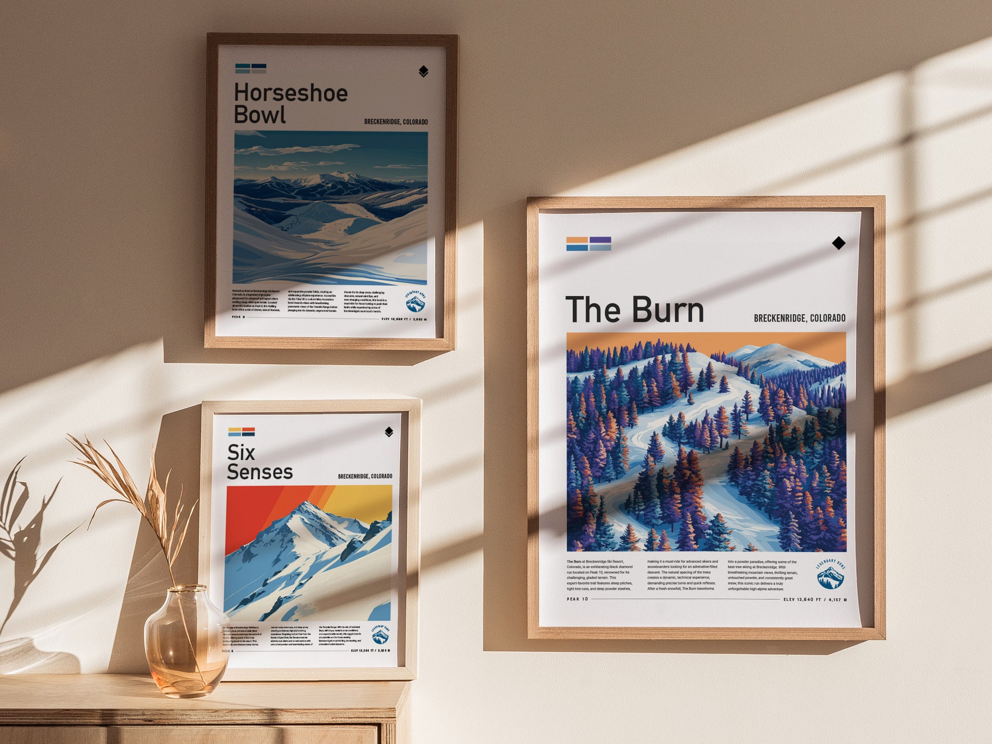 Three framed ski run posters are displayed on a wall, featuring The Burn, Horseshoe Bowl, and Six Senses from Breckenridge, Colorado. Sunlight casts shadows across the scene.