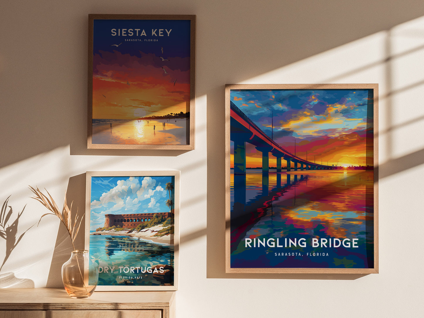 Three framed posters hang on a sunlit wall. The largest shows a vibrant sunset over the Ringling Bridge, reflecting in the water. The others depict a sunset at Siesta Key and a scenic view of Dry Tortugas with bright skies.