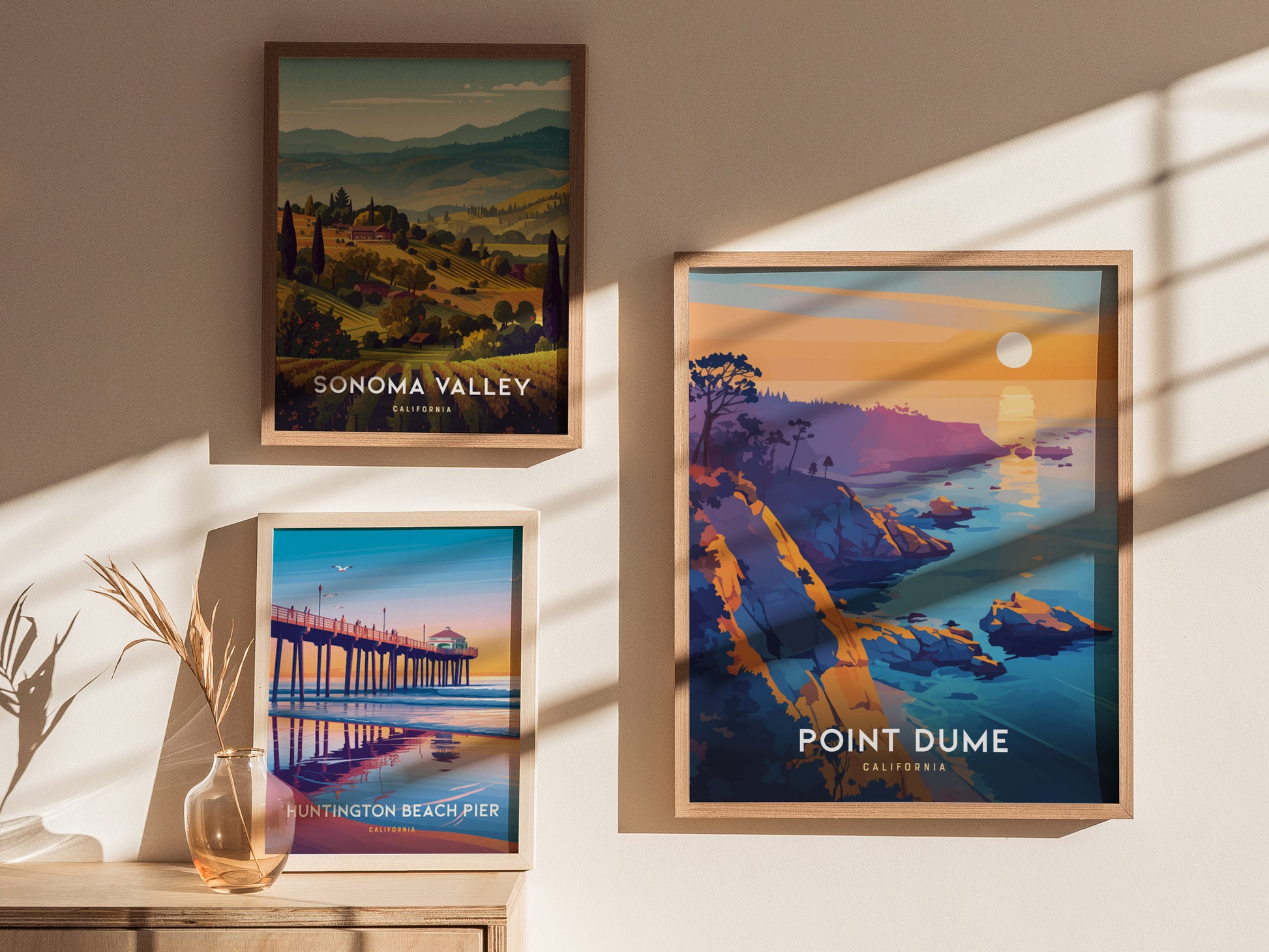 Three framed posters hang on a sunlit wall, featuring scenic landscapes of Sonoma Valley, Point Dume, and Huntington Beach Pier, with a vase of dried plants below.