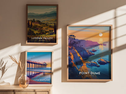 Three framed posters hang on a sunlit wall, featuring scenic landscapes of Sonoma Valley, Point Dume, and Huntington Beach Pier, with a vase of dried plants below.