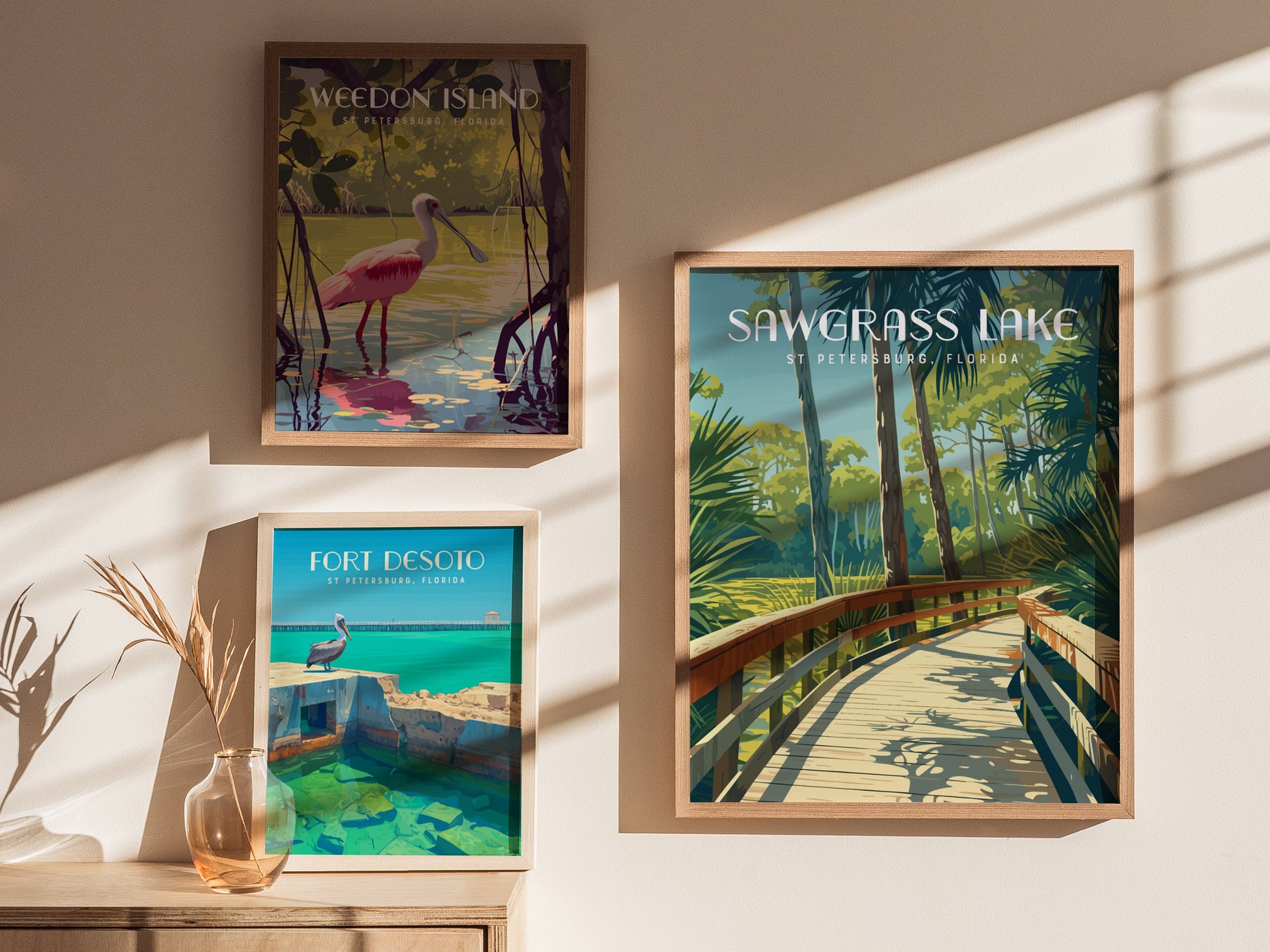 Three framed posters are displayed on a sunlit wall, featuring scenic illustrations of Sawgrass Lake, Weedon Island, and Fort Desoto, each capturing vibrant natural landscapes.
