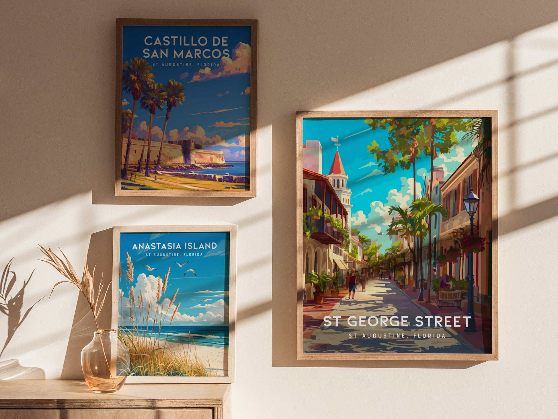 Three framed posters hang on a sunlit wall, depicting St. Augustine locations: Castillo de San Marcos, Anastasia Island, and St. George Street, showcasing vibrant travel scenes.