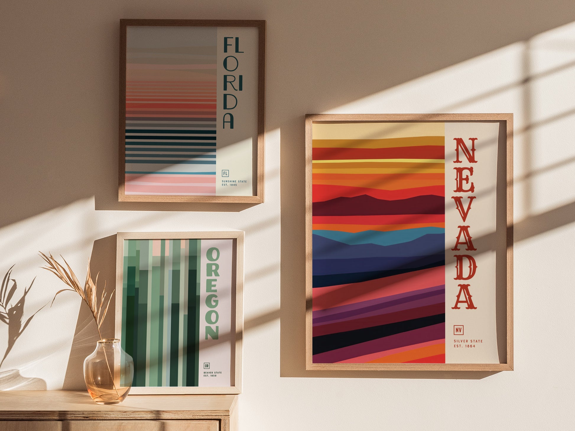 Three framed minimalist posters are displayed on a wall, each representing Florida, Oregon, and Nevada with abstract horizontal and vertical lines in various colors. Sunlight casts soft shadows.