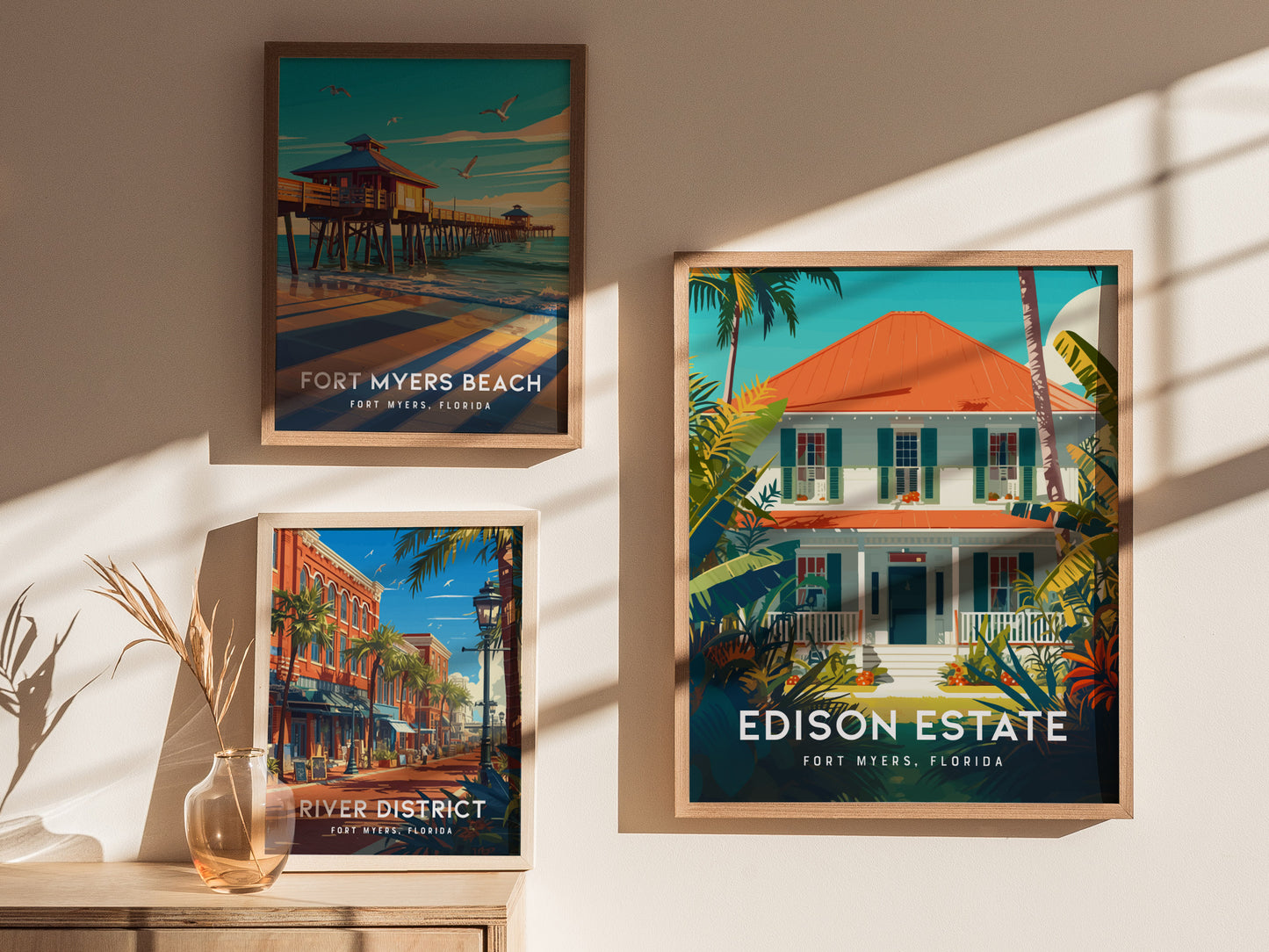 Three framed posters on a wall depict Fort Myers Beach, the River District, and Edison Estate in vibrant colors, with sunlight casting shadows across them.