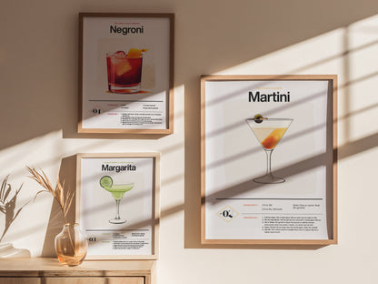 Three framed cocktail posters are displayed on a beige wall, featuring Martini, Negroni, and Margarita recipes. Sunlight casts shadows, creating a warm and inviting atmosphere.
