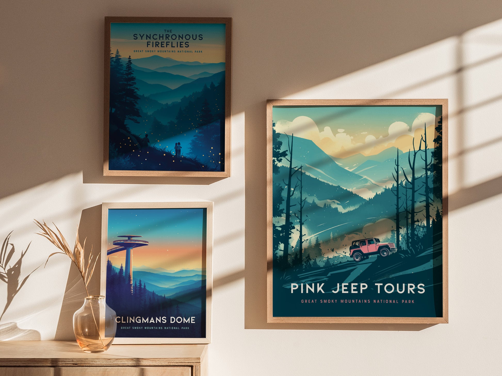 Three framed posters are displayed on a sunlit wall. The largest features a pink jeep navigating a mountainous landscape. The other two depict scenes from the Great Smoky Mountains National Park with themes of fireflies and Clingmans Dome.