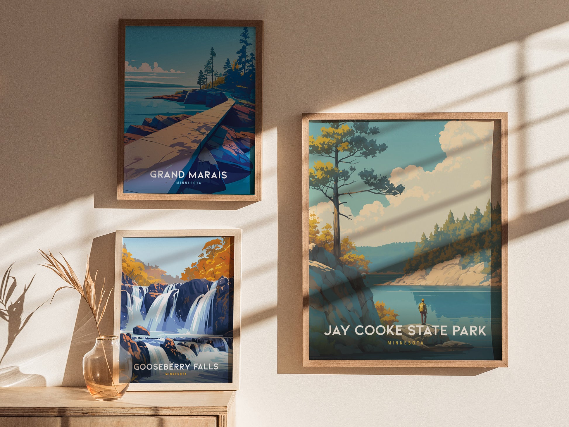 Three framed posters on a wall depict scenic landscapes: Grand Marais, Gooseberry Falls, and Jay Cooke State Park. Sunlight casts shadows, and a vase with dried plants sits below.