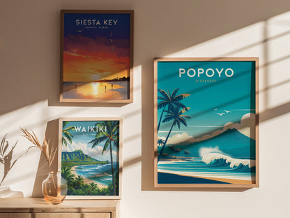 Three framed travel posters are displayed on a wall, featuring Siesta Key, Waikiki, and Popoyo, with vibrant beach scenes and palm trees. A vase with dried plants is nearby.