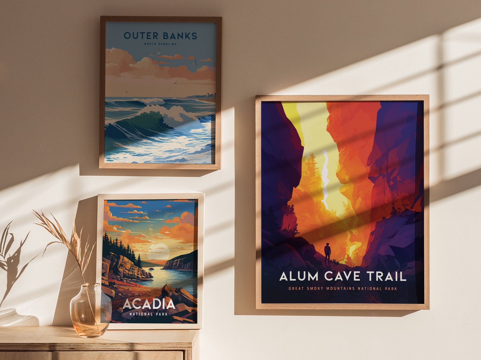 Three framed posters hang on a sunlit wall: Outer Banks with ocean waves, Acadia featuring a sunset over cliffs, and Alum Cave Trail depicting a vibrant, colorful canyon scene.