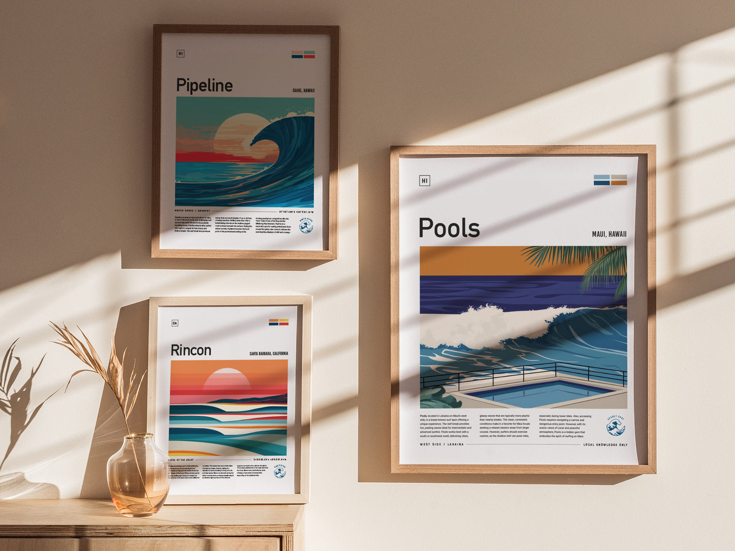 Three framed surf posters are displayed on a beige wall, featuring Pipeline, Rincon, and Pools designs. Sunlight casts shadows, and a vase with dried plants sits below.