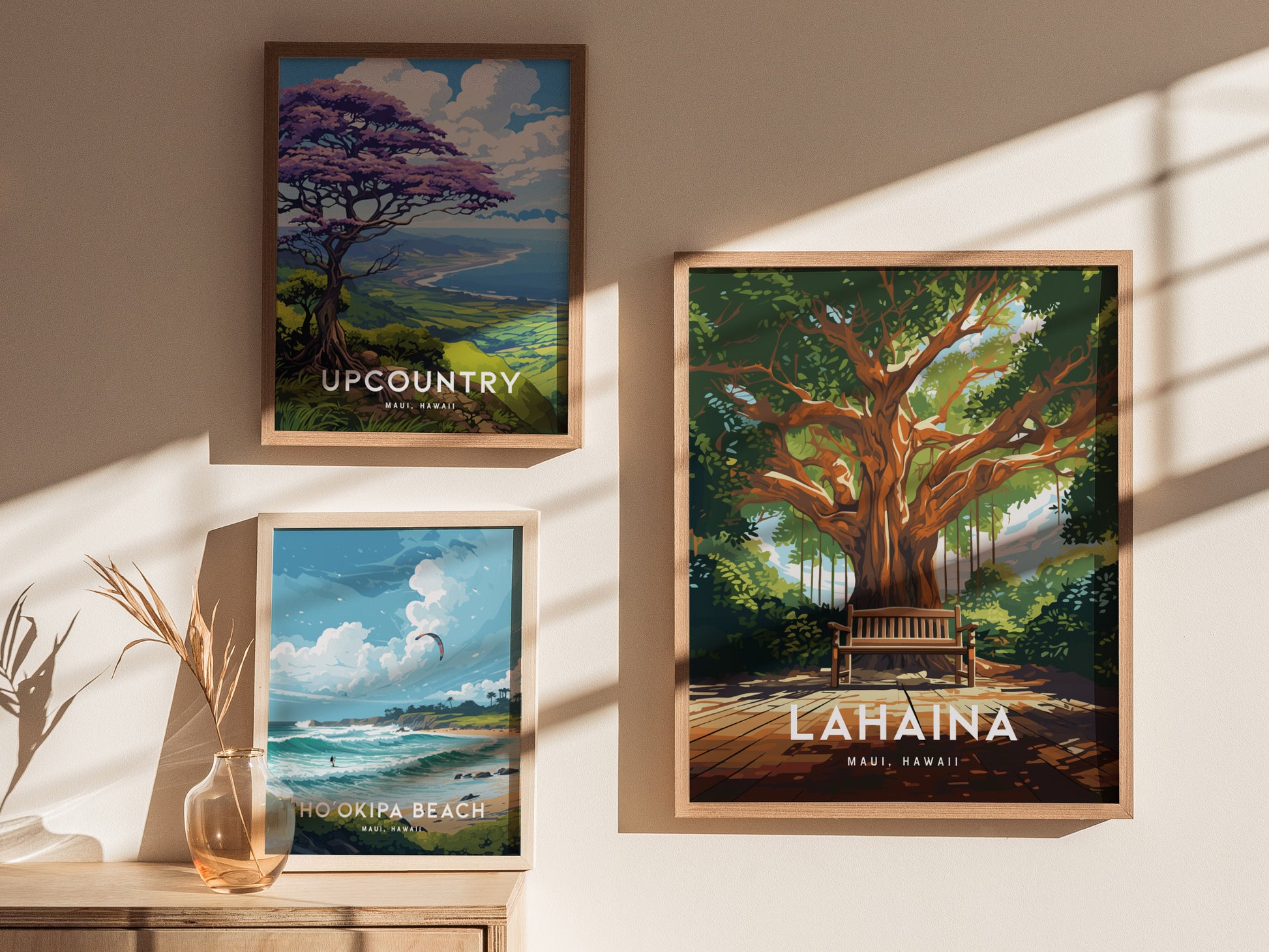 Three framed posters are displayed on a sunlit wall, featuring scenic artwork of Upcountry Maui, Ho‘okipa Beach, and the Lahaina Banyan Tree with a bench beneath its canopy.