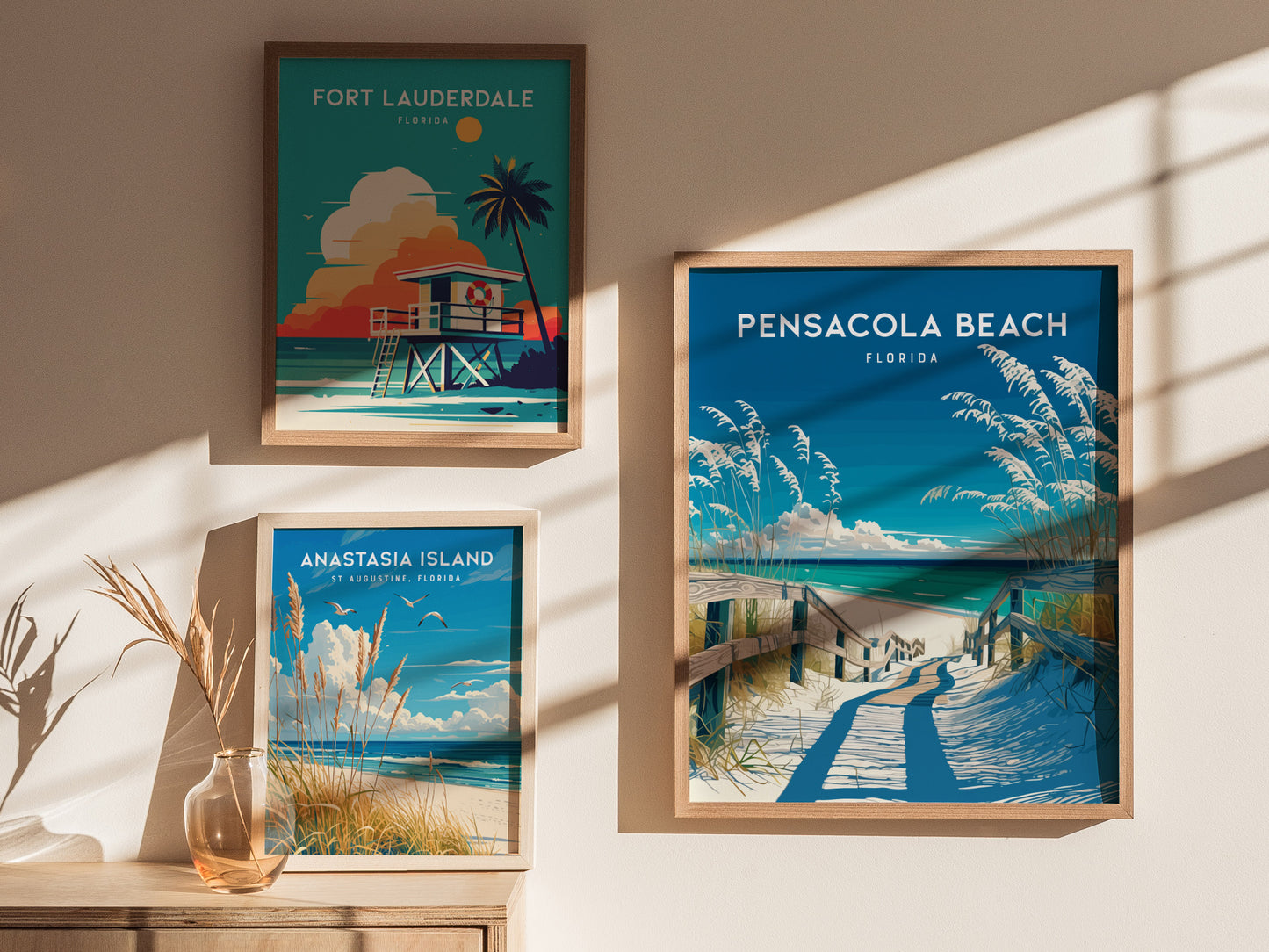 Three framed posters depict Florida beaches: Pensacola Beach, Fort Lauderdale, and Anastasia Island. They are displayed on a sunlit wall beside a vase with dried grasses.