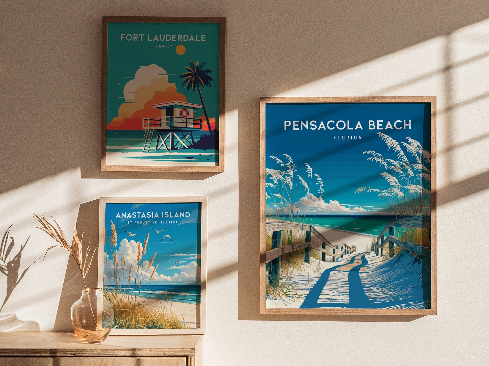 Three framed posters depict Florida beaches: Pensacola Beach, Fort Lauderdale, and Anastasia Island. They are displayed on a sunlit wall beside a vase with dried grasses.