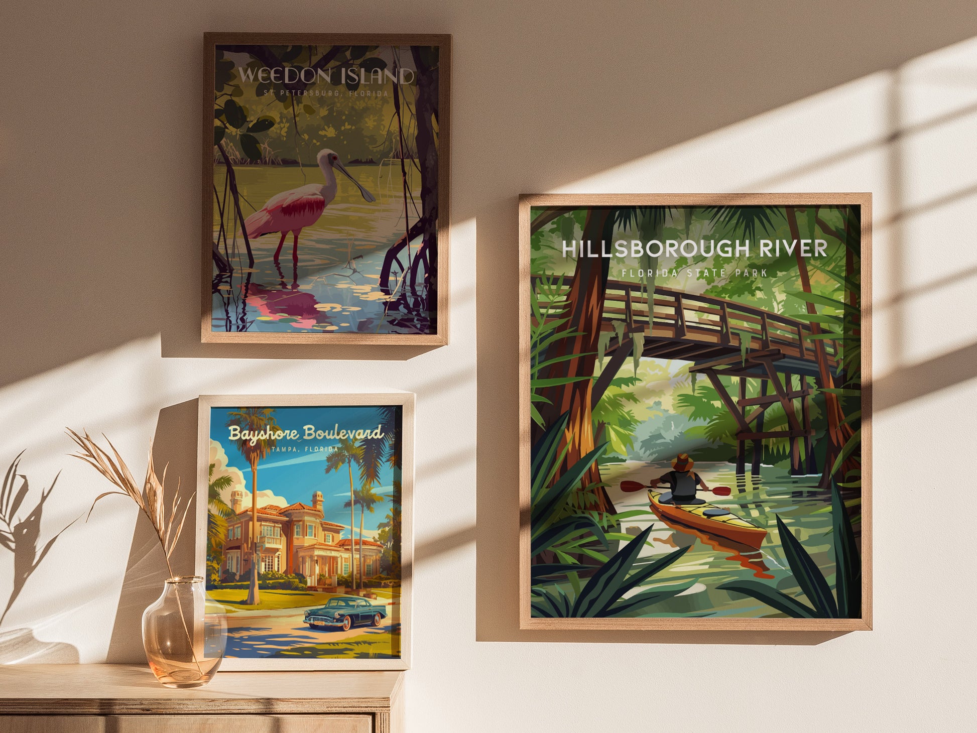 Three framed posters are displayed on a wall. The largest features a kayaker on a river under a wooden bridge, surrounded by lush greenery. Shadows and sunlight add depth.