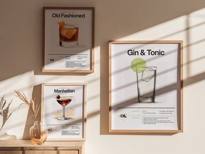Three framed cocktail posters featuring Old Fashioned, Gin & Tonic, and Manhattan drinks are displayed on a sunlit wall above a wooden surface with a small vase.