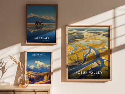 Three framed posters hang on a wall, displaying scenic illustrations of Lake Clark, Kobuk Valley, and Mount Denali. Sunlight casts soft shadows across the artwork and wooden surface.