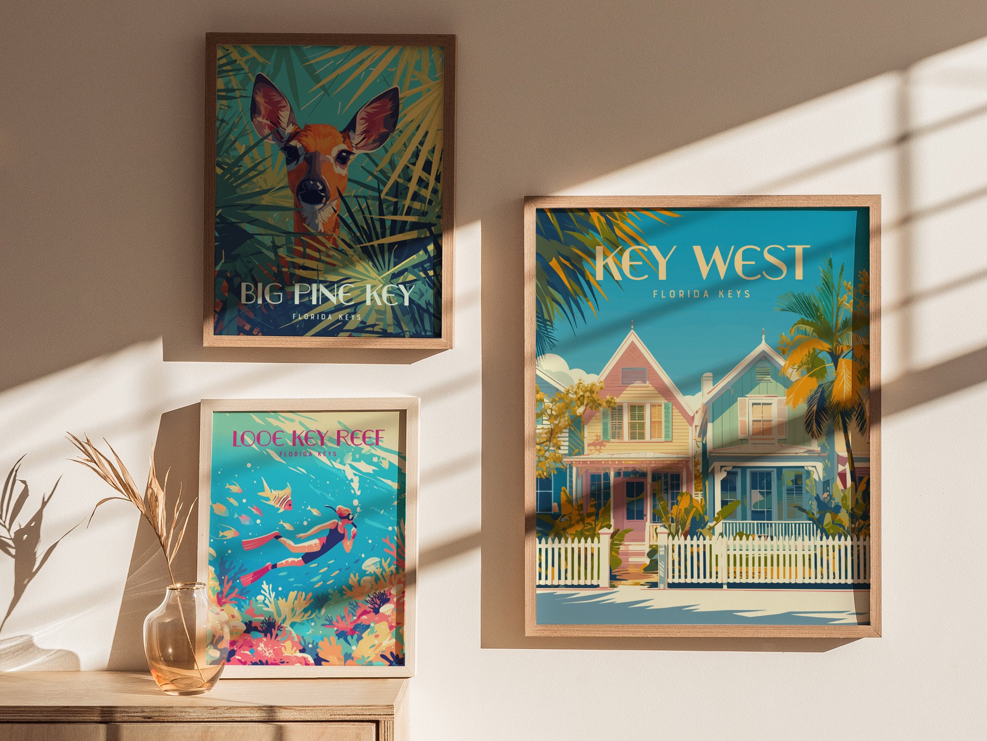 Three framed posters hang on a sunlit wall: a deer in foliage, pastel Key West houses, and a diver with colorful coral. A glass vase with dried stems sits below.