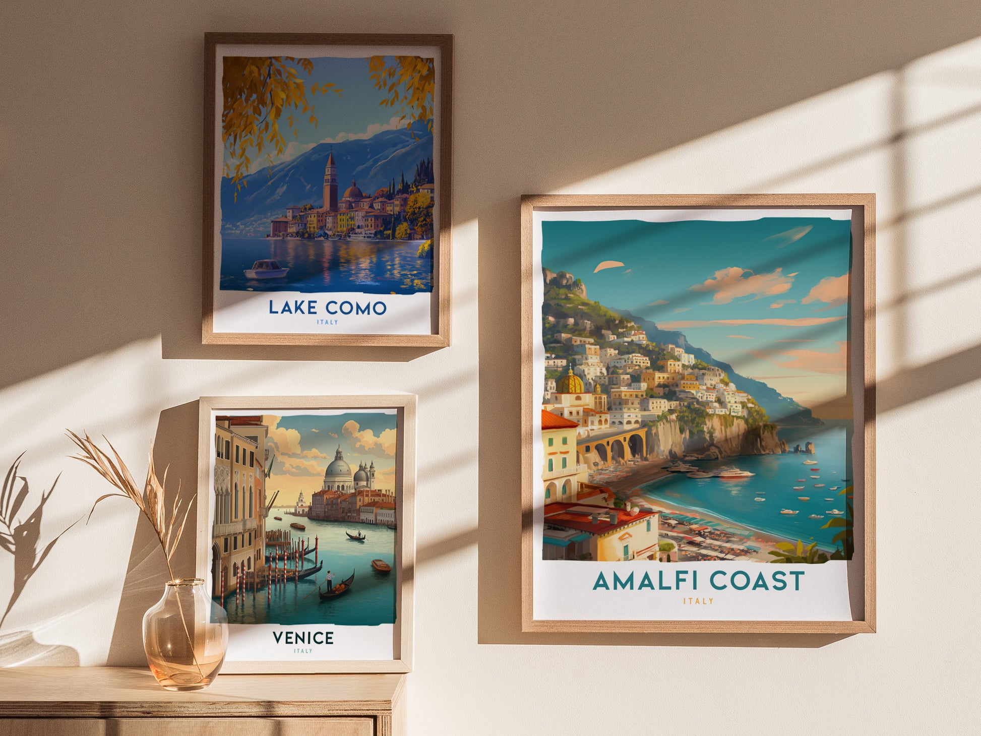 Three framed travel prints are displayed on a sunlit wall, featuring scenic views of Lake Como, Venice, and the Amalfi Coast in Italy, with a vase on a wooden surface.