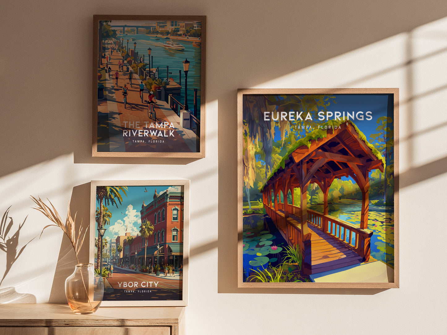 Three framed posters hang on a wall, depicting vibrant illustrations of Tampa, Florida: the Tampa Riverwalk, Ybor City, and Eureka Springs Park with a covered wooden bridge.