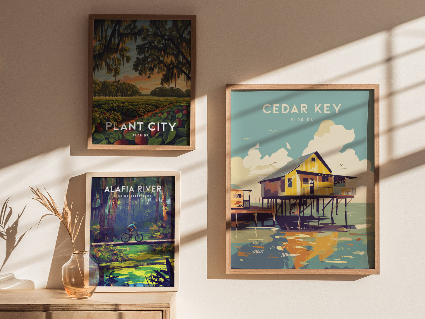 Framed posters of Cedar Key, Plant City, and Alafia River are displayed on a sunlit wall. A vase with dried plants sits on a wooden surface below.