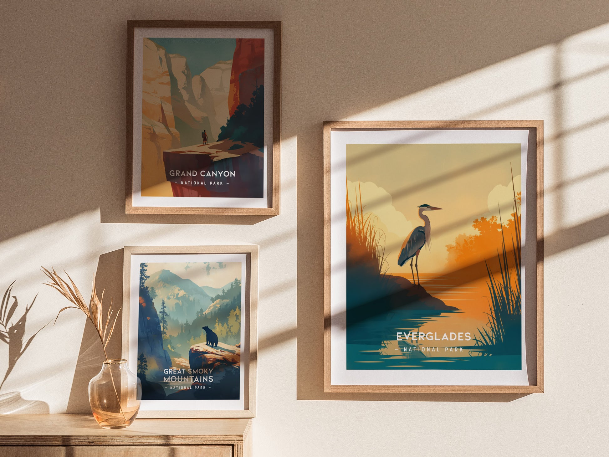 Three framed posters on a sunlit wall depict Grand Canyon, Great Smoky Mountains, and Everglades National Parks, featuring colorful landscapes and wildlife silhouettes. A vase with dried plants sits below.